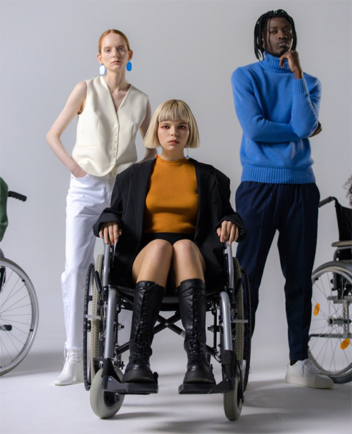 Woman sitting in wheelchair and 2 people of different nationalities standing behind her.